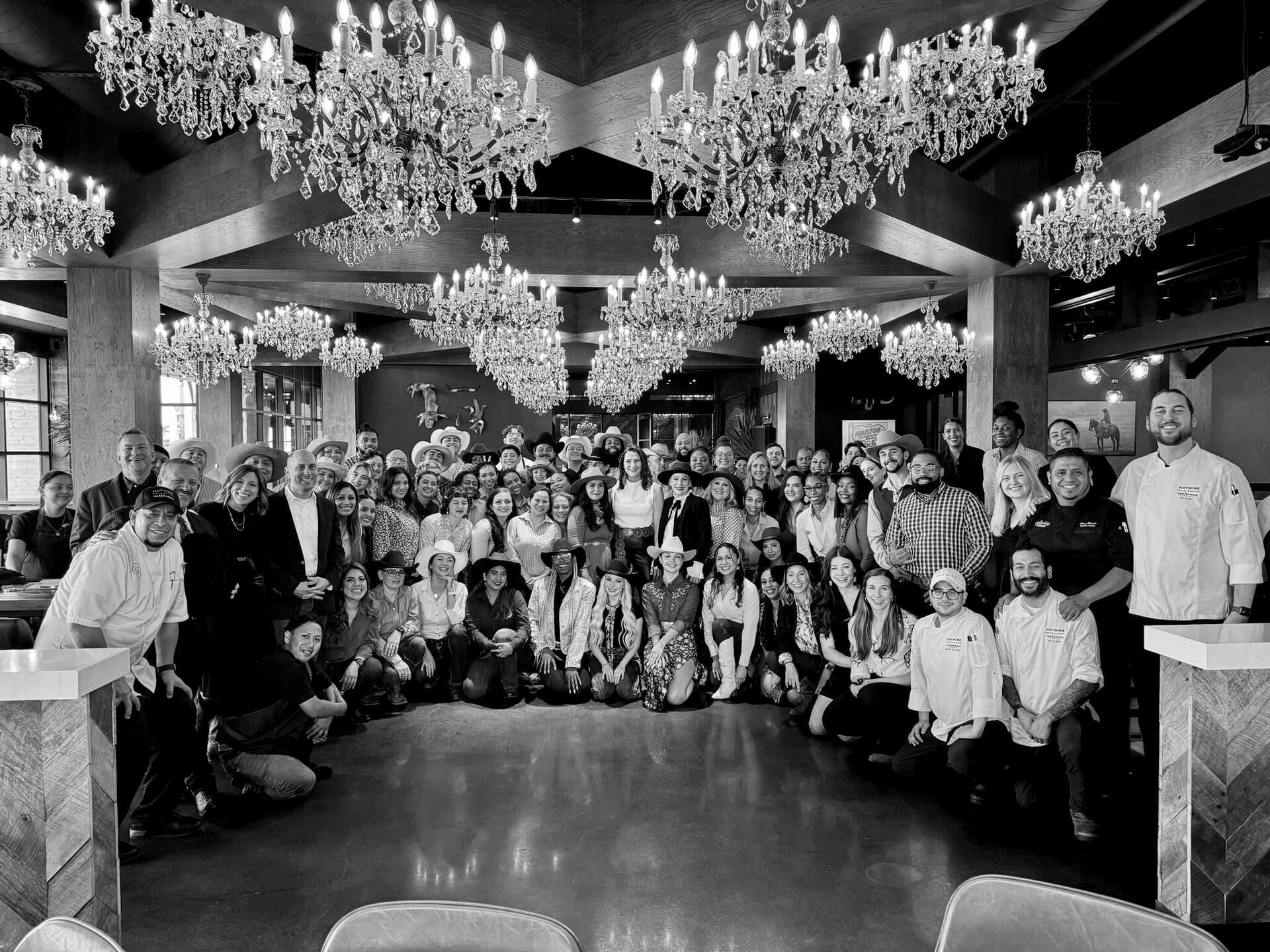 The full opening team - chefs in whites, servers, and leadership - gathered under crystal chandeliers in the timber-beamed dining room at Haywire's Memorial opening