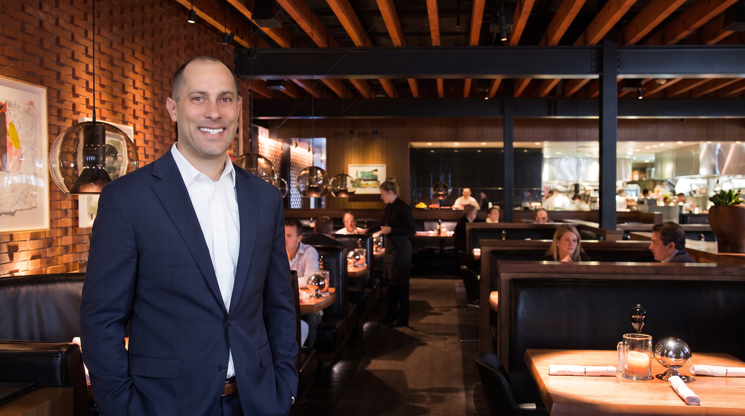 Jon Peck in a navy suit standing in a warm brick-walled dining room, with leather booths, glass pendant lights, and an open kitchen behind him