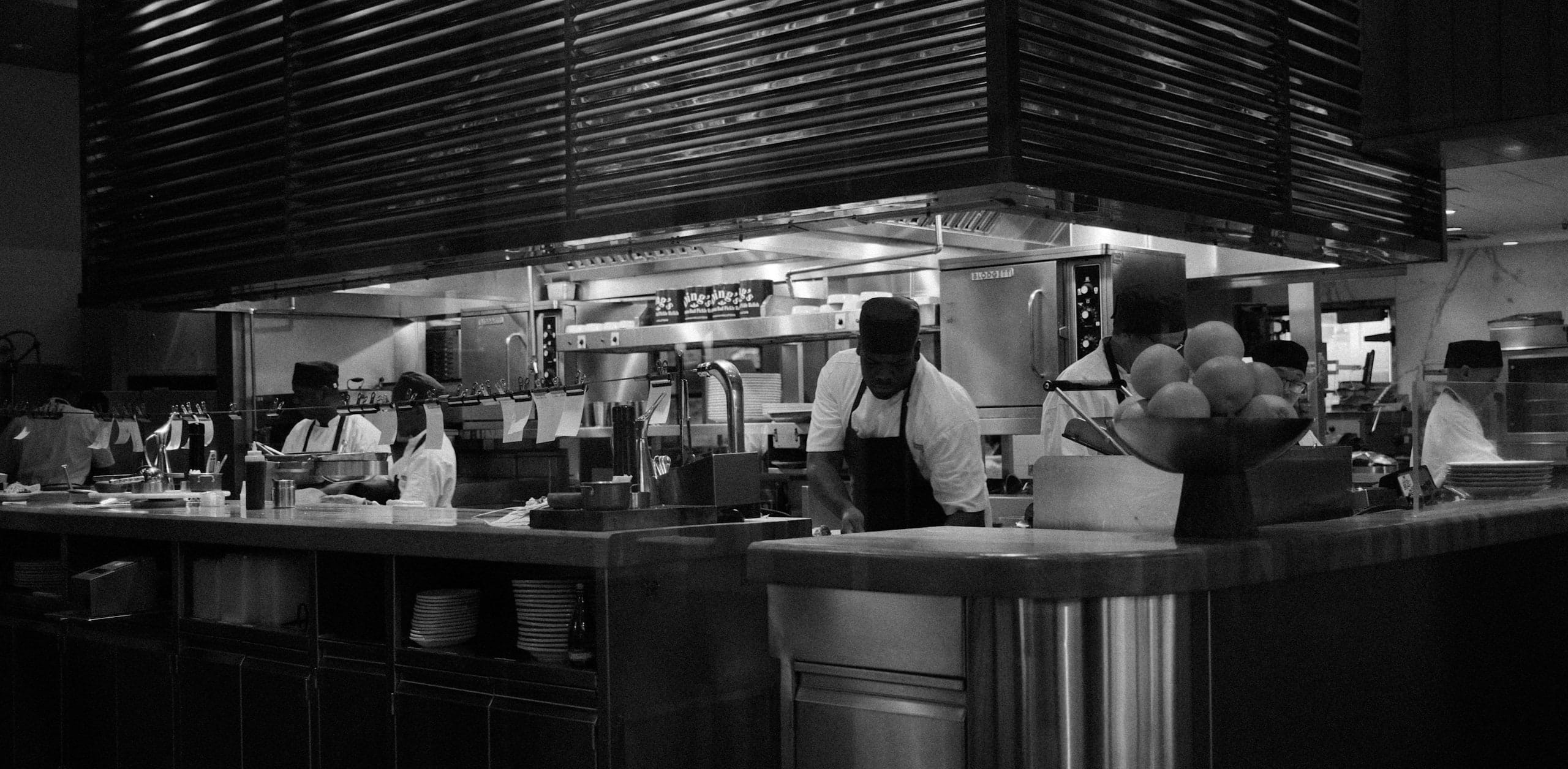 An open kitchen mid-service: cooks working behind the expo pass under a slatted hood