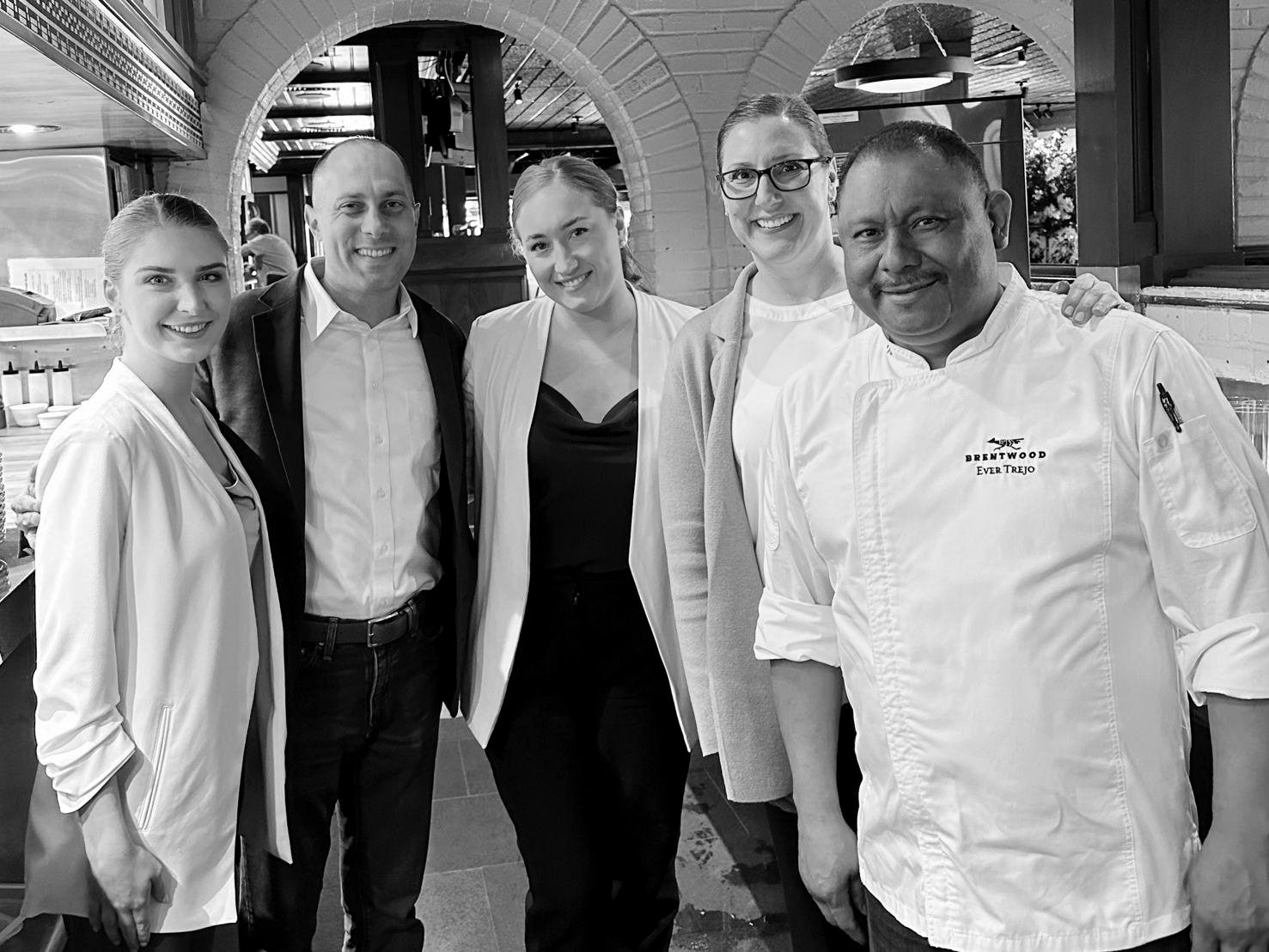 Five of the Brentwood leadership team - managers, the chef in whites - smiling in front of the restaurant's brick archways and open kitchen