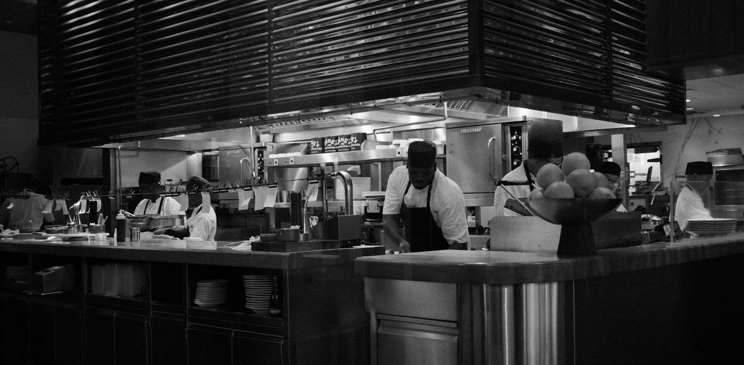 A black-and-white photograph of a working open kitchen, cooks moving behind an expo pass under a slatted hood